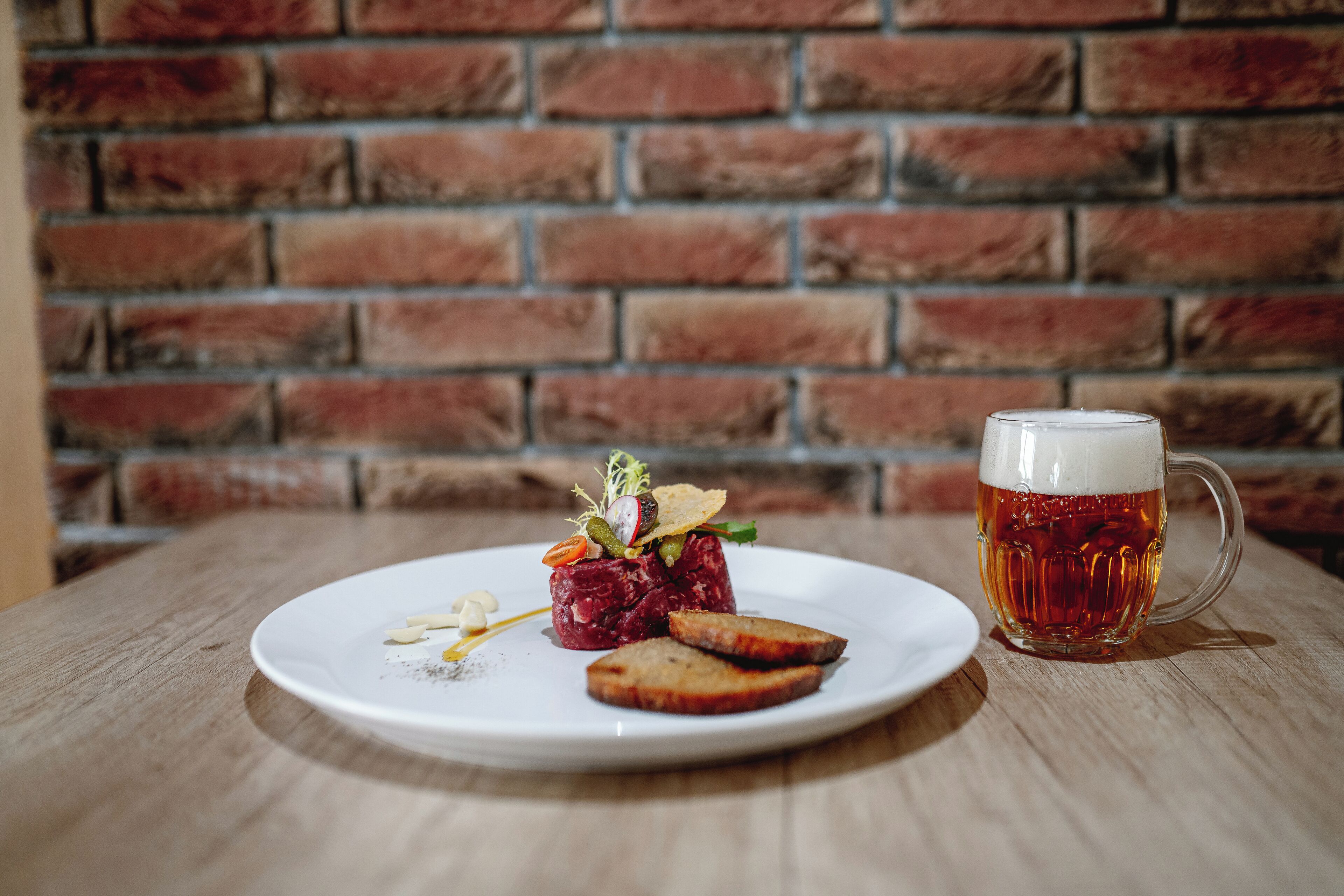 Un plato con tártara de carne, tostadas y una jarra de cerveza sobre una mesa de madera frente a una pared de ladrillo.