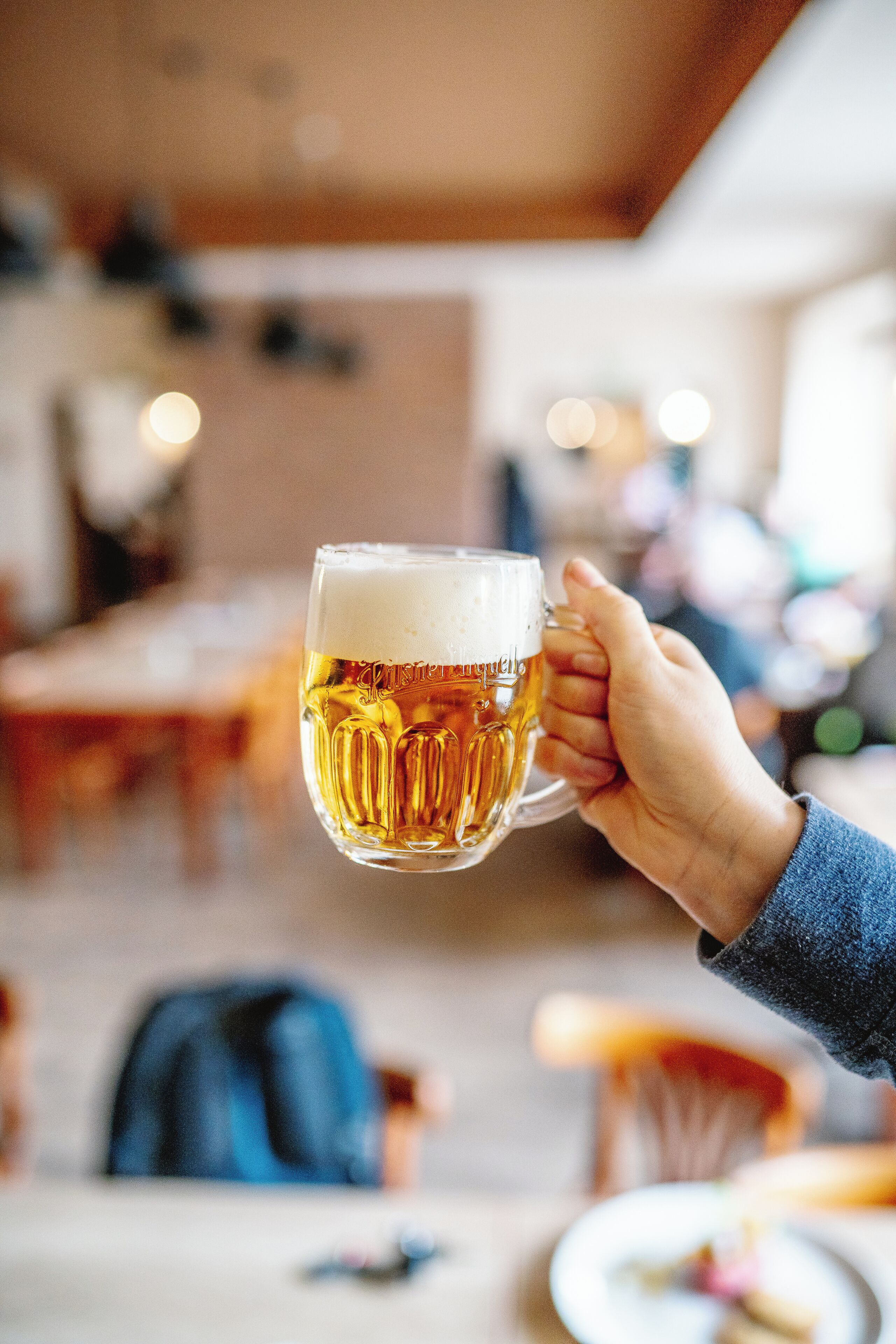 A hand holding a glass of beer with a blurred background of a dining area.