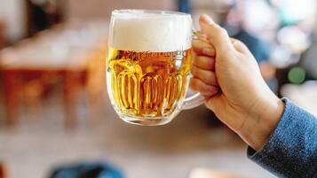 A hand holding a glass of beer with a blurred background of a dining area.