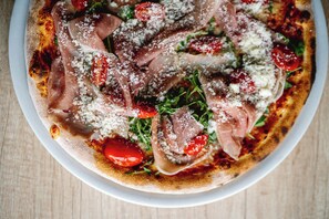 A pizza with prosciutto, tomatoes, and cheese on a white plate. - Hotel Prácheň (Horazdovice)