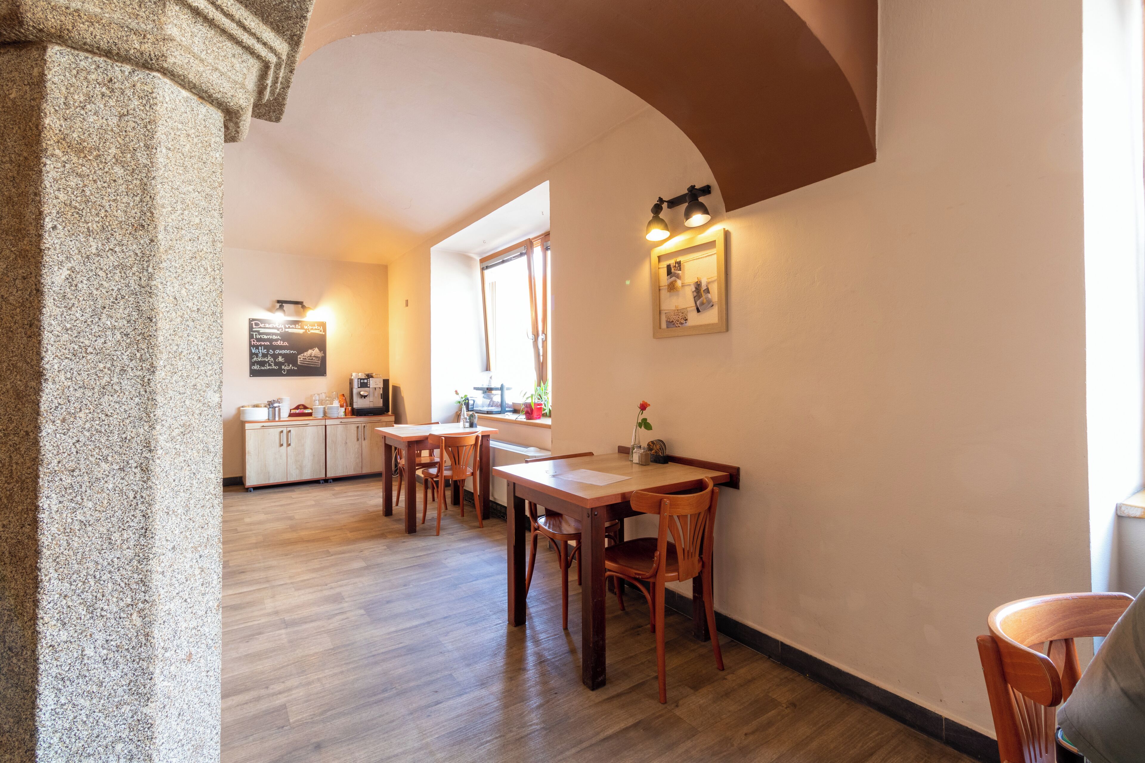 A dining area with wooden tables and chairs, a stone column, and a kitchen in the background.