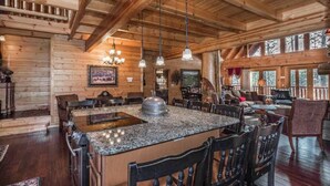 Una cocina amplia con encimeras de granito, armarios de madera y una isla grande. Hay varias lámparas colgantes y un candelabro. La cocina está adyacente a un comedor con varias sillas y una mesa.
