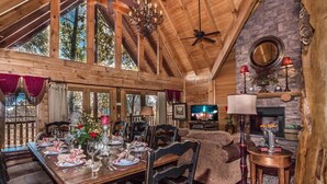 Interior rústico de una cabaña de madera con una mesa de comedor dispuesta para una comida, una chimenea y un televisor al fondo.