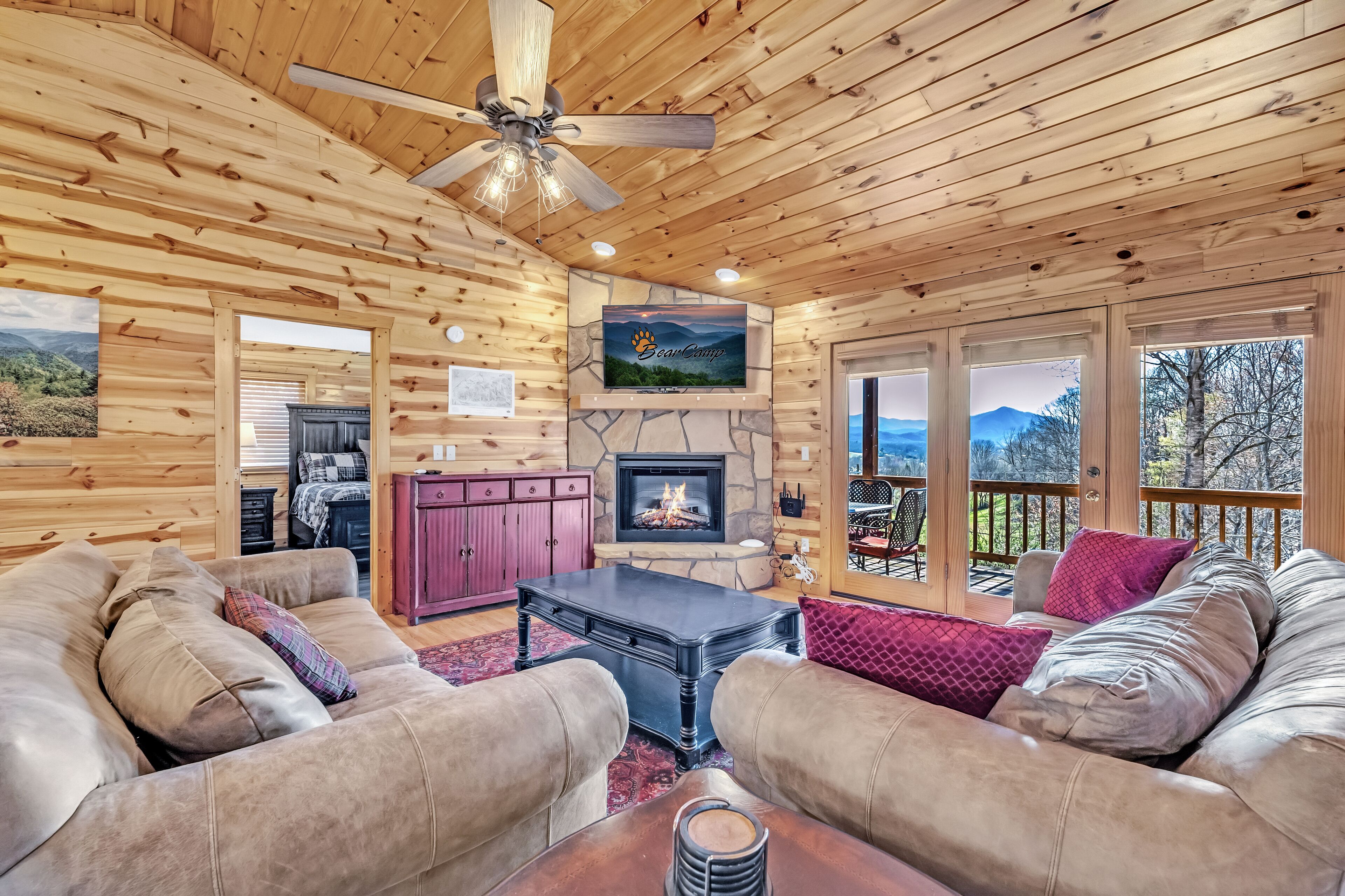 A cozy log cabin interior with a stone fireplace, leather sofas, and a large window overlooking a scenic landscape.