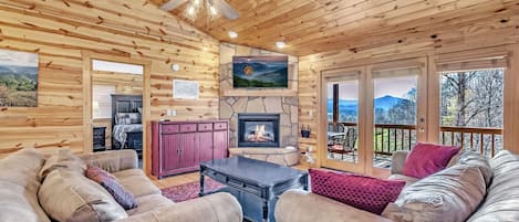 A cozy log cabin interior with a stone fireplace, leather sofas, and a large window overlooking a scenic landscape.