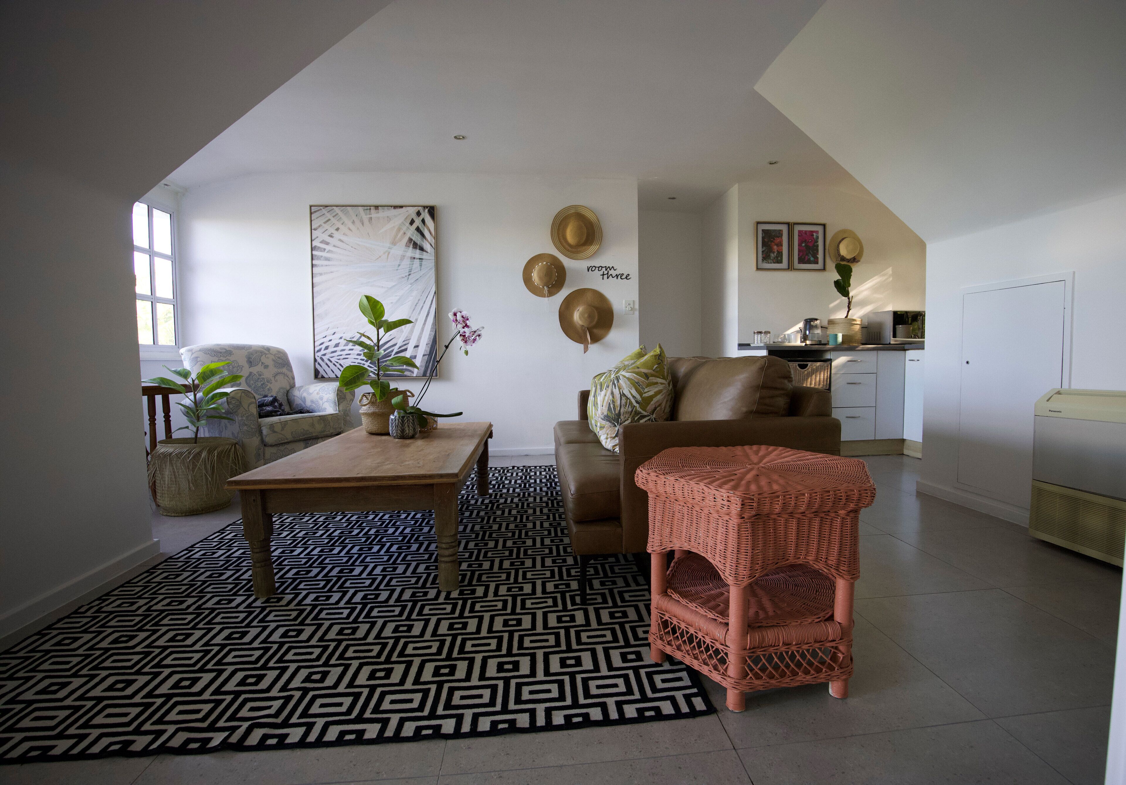 A living room with a geometric patterned rug, a wooden coffee table, a brown sofa, a wicker chair, and a kitchen area in the background.