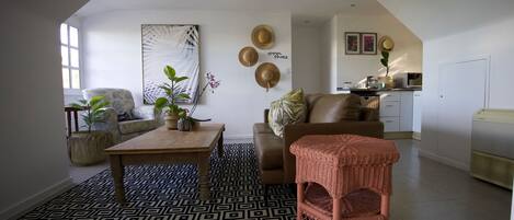 A living room with a geometric patterned rug, a wooden coffee table, a brown sofa, a wicker chair, and a kitchen area in the background.