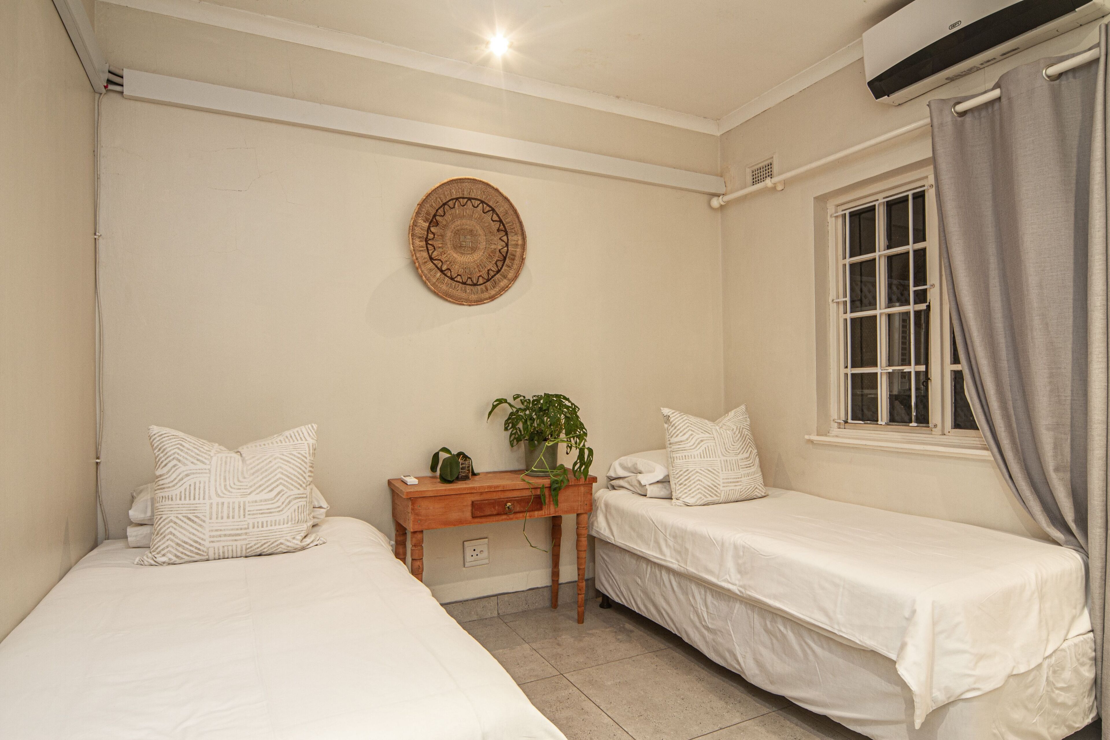 A bedroom with two single beds, a wooden nightstand with a plant, a wall-mounted circular decoration, and a window with curtains.
