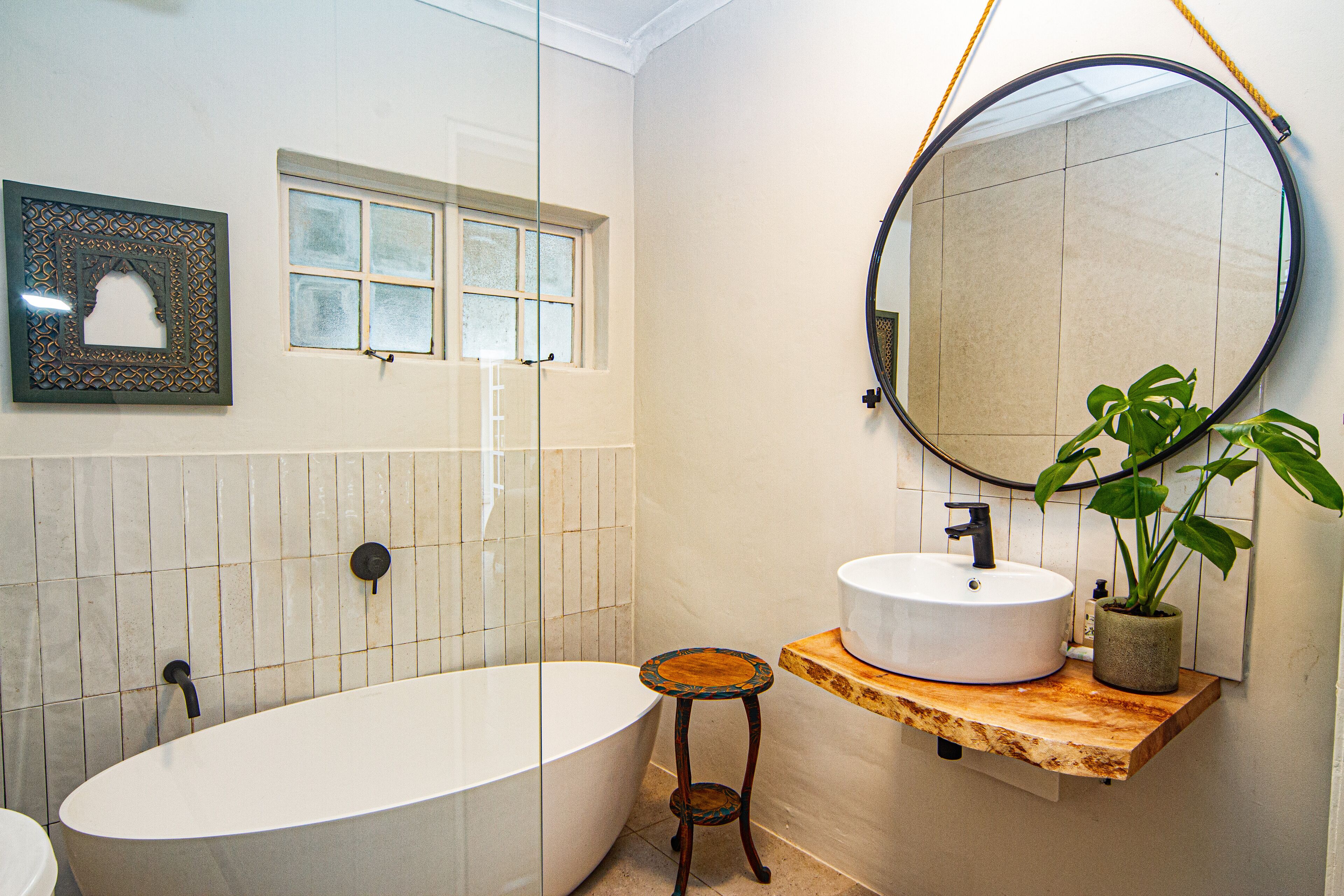A bathroom with a freestanding bathtub, a round mirror, a sink on a wooden shelf, a potted plant, and a framed picture on the wall.