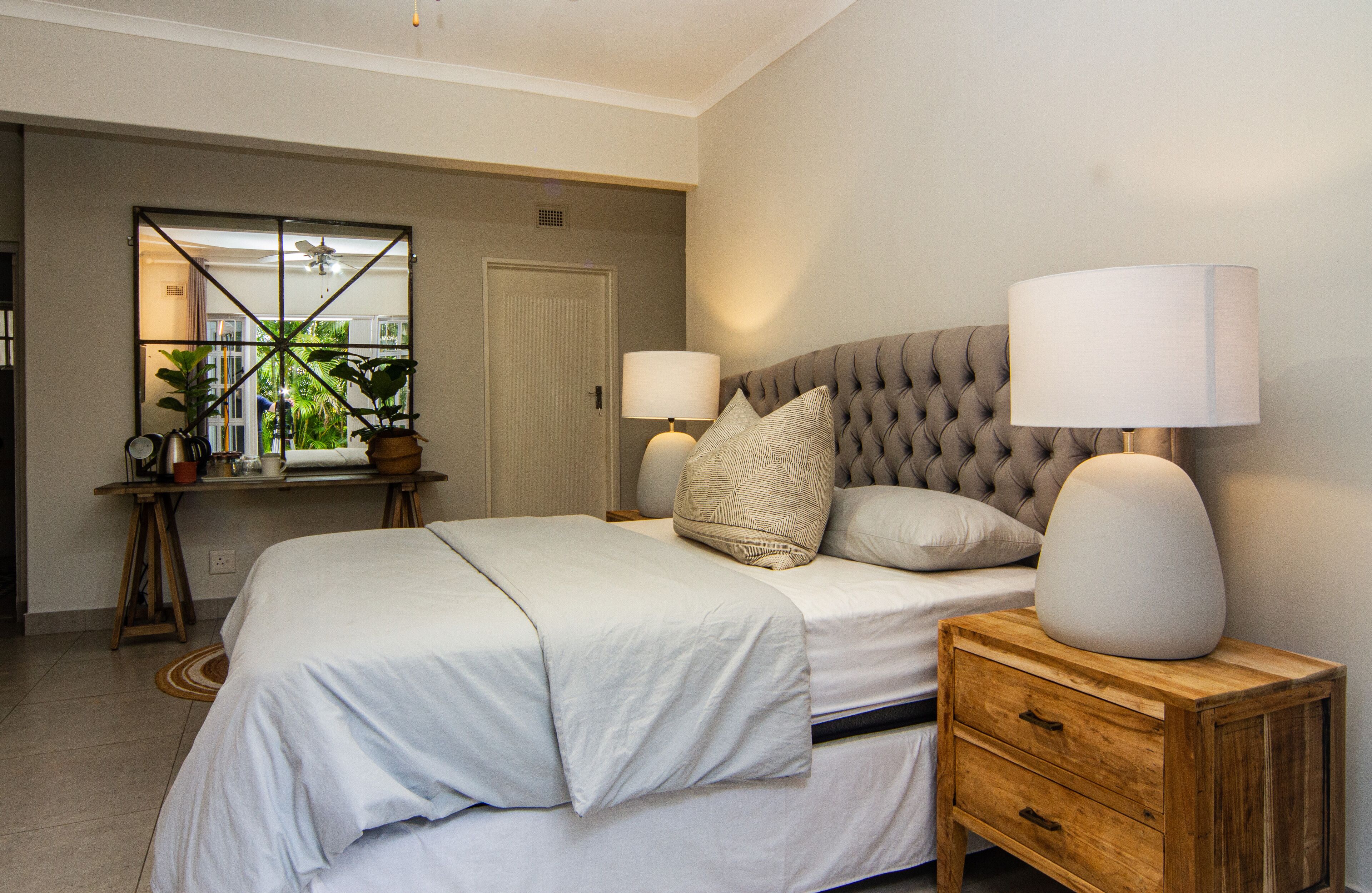 Un dormitorio con una cama grande, dos mesitas de noche con lámparas, un espejo y vistas al exterior con vegetación.
