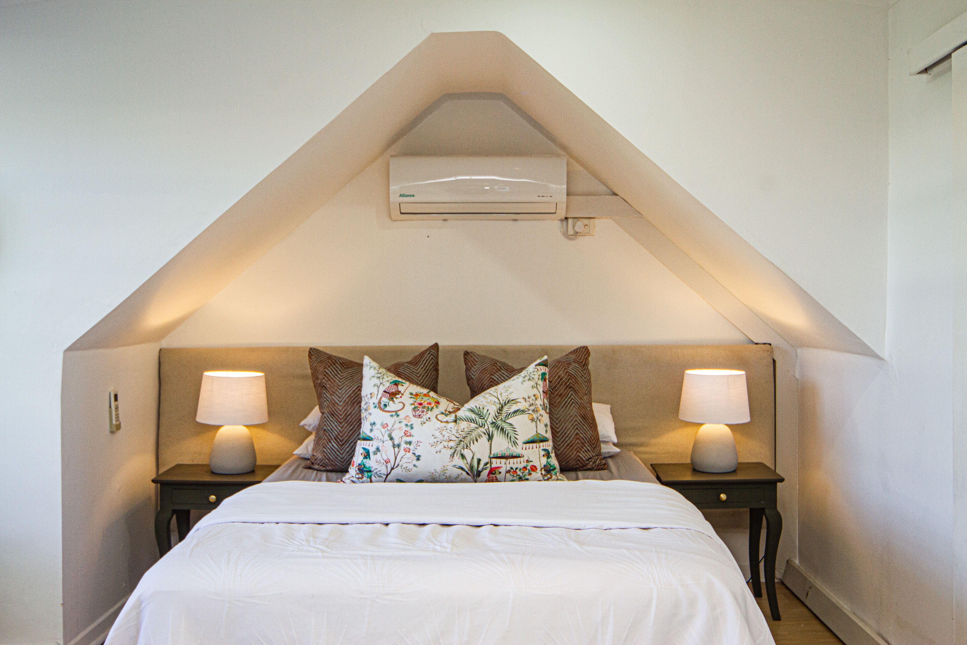 Un dormitorio con techo inclinado, una cama con ropa blanca, dos lámparas de noche y almohadones decorativos.