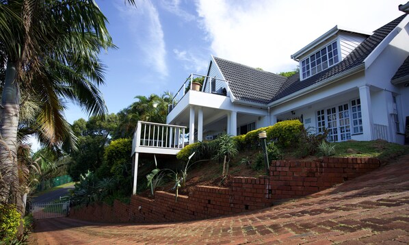 A two-story house with a balcony, surrounded by lush greenery and a brick retaining wall.