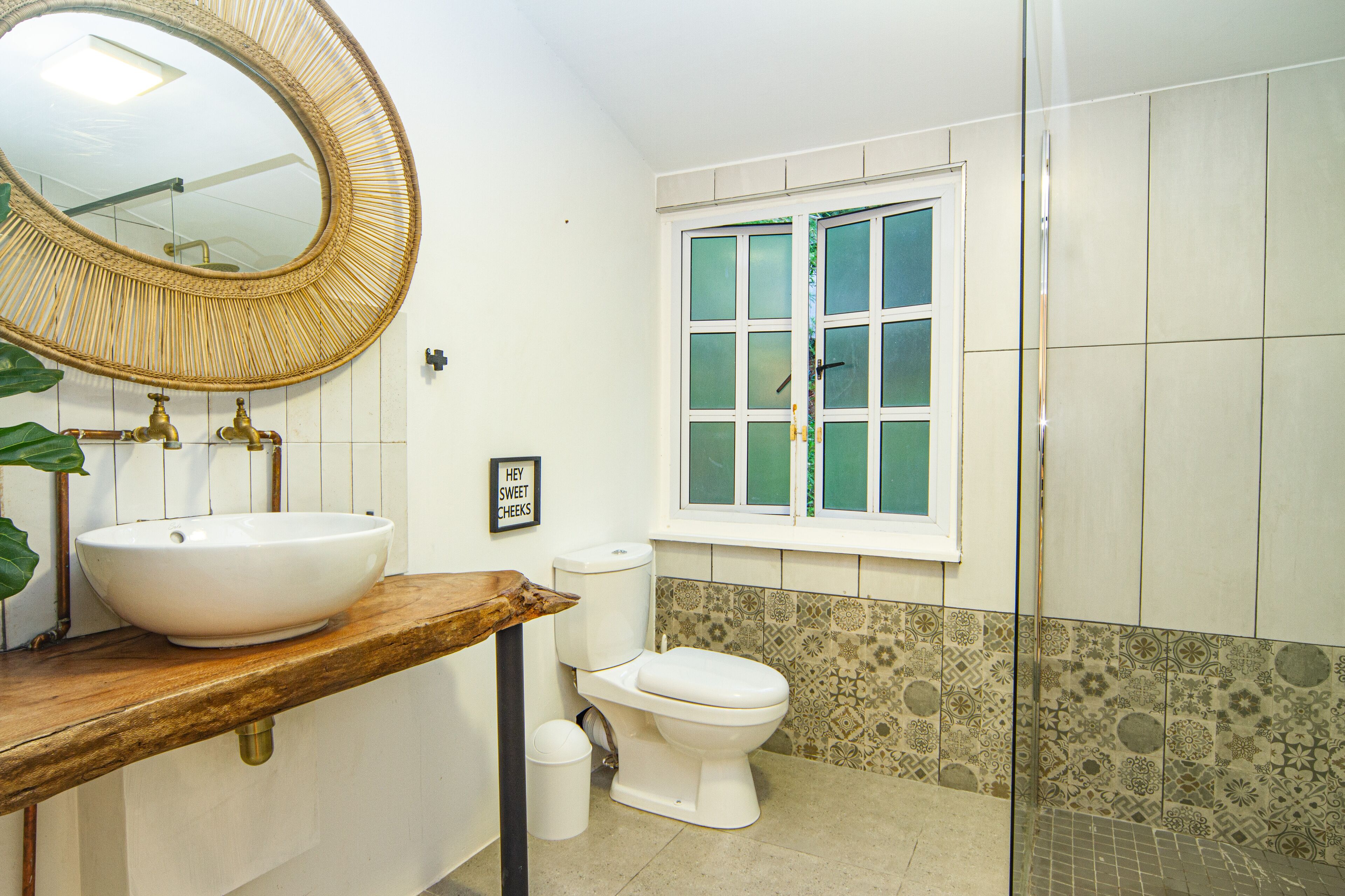 A bathroom with a round mirror, a wooden countertop, a white sink, a toilet, and a window with frosted glass panes.
