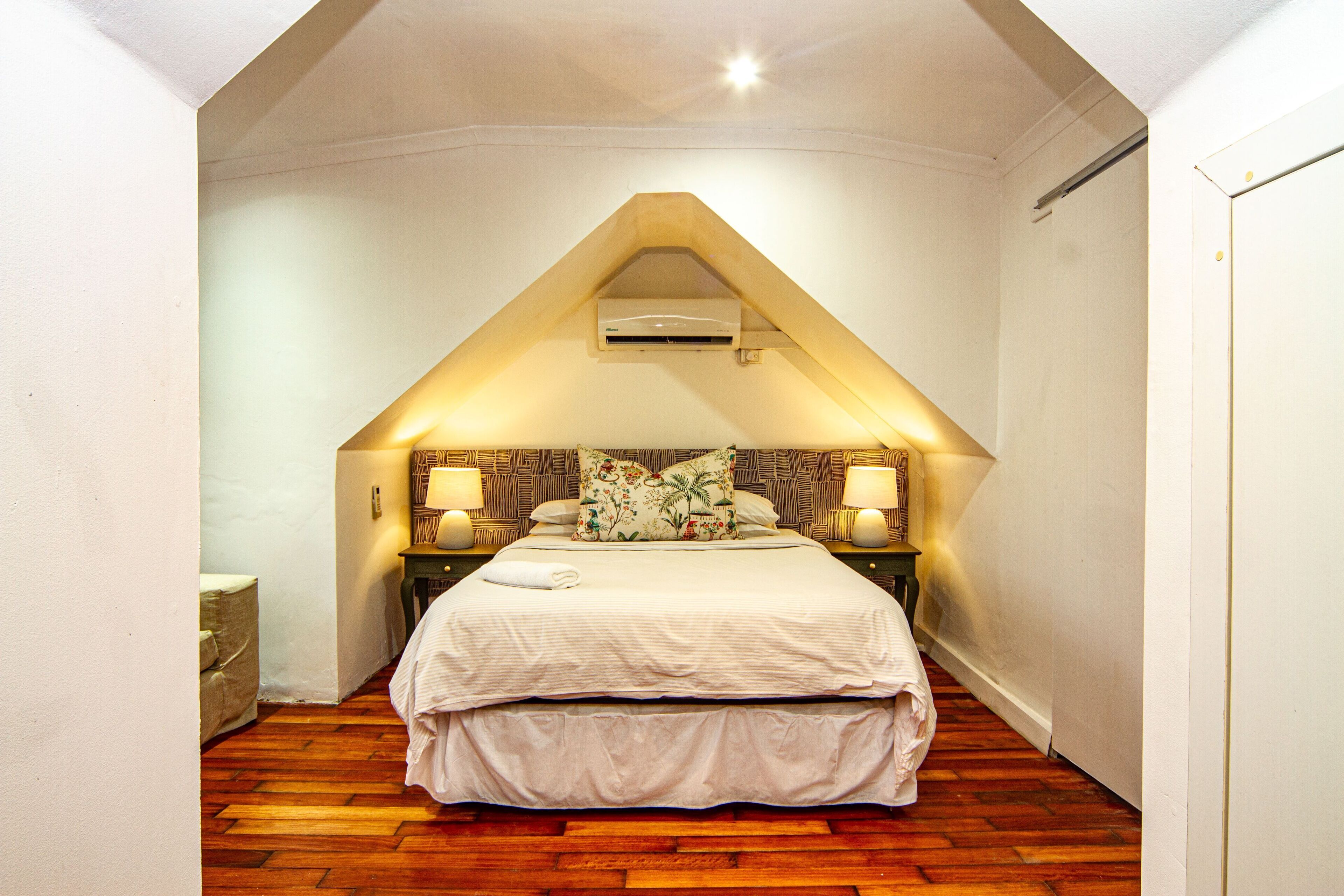 A bedroom with a sloped ceiling, a bed with white bedding, two bedside lamps, and an air conditioning unit.