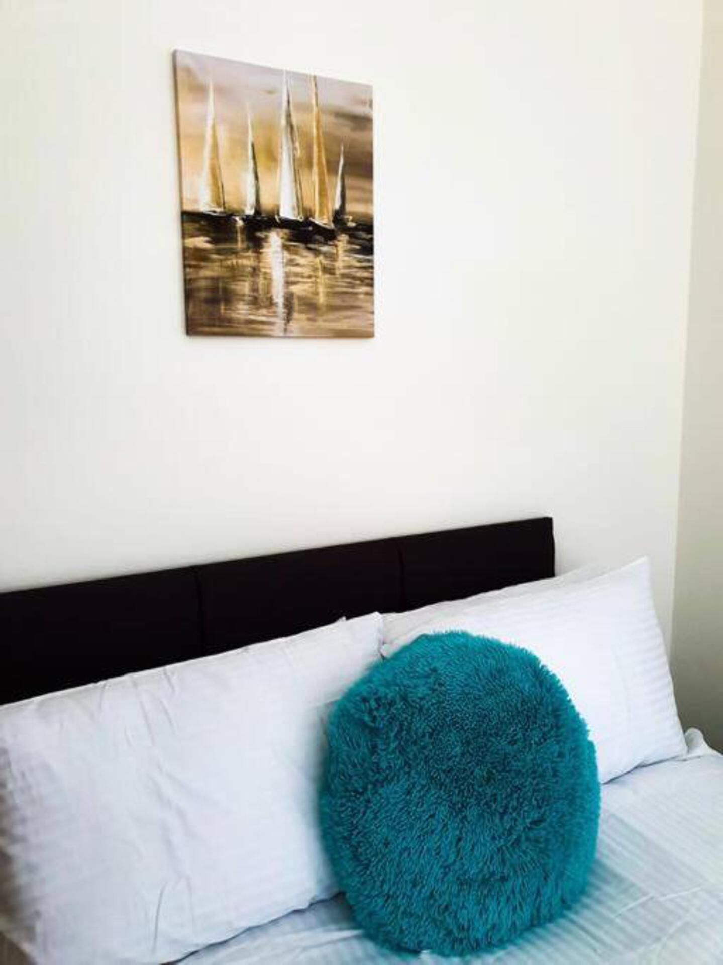 Un dormitorio con una cama, almohadas blancas, un cojín azul turquesa y un cuadro con barcos a vela en la pared.