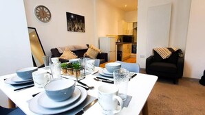 A dining area with a white table set for four, including plates, bowls, glasses, and cutlery. There is a black couch in the background, a clock on the wall, and a framed picture above the couch.