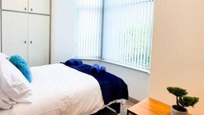 A bedroom with a bed, a wardrobe, a bedside table with a plant, and a window with blinds.