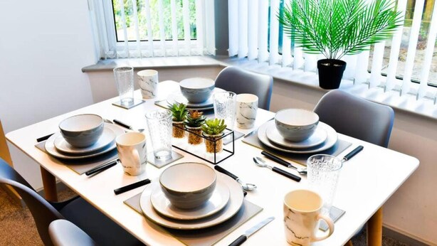 A dining table set with plates, bowls, and cutlery, surrounded by chairs, with a potted plant and a window with blinds in the background.