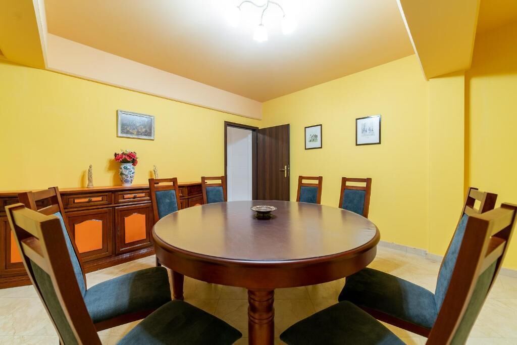 A dining area with a round table and six chairs, yellow walls, and a wooden cabinet with a vase of flowers.