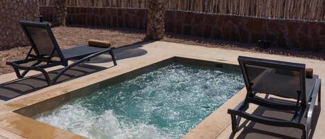 A pool area with two lounge chairs, a small hot tub, and a thatched roof structure.