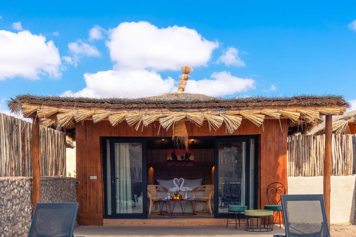 A pool area with a thatched-roof structure, lounge chairs, and a table with fruit.