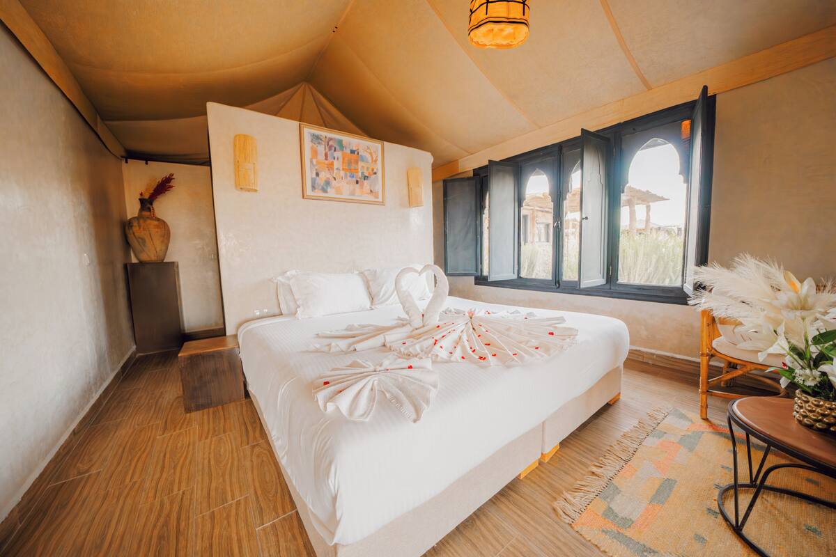 A spacious bedroom with a large bed, a wooden floor, a window with a view, and a small table with a vase.
