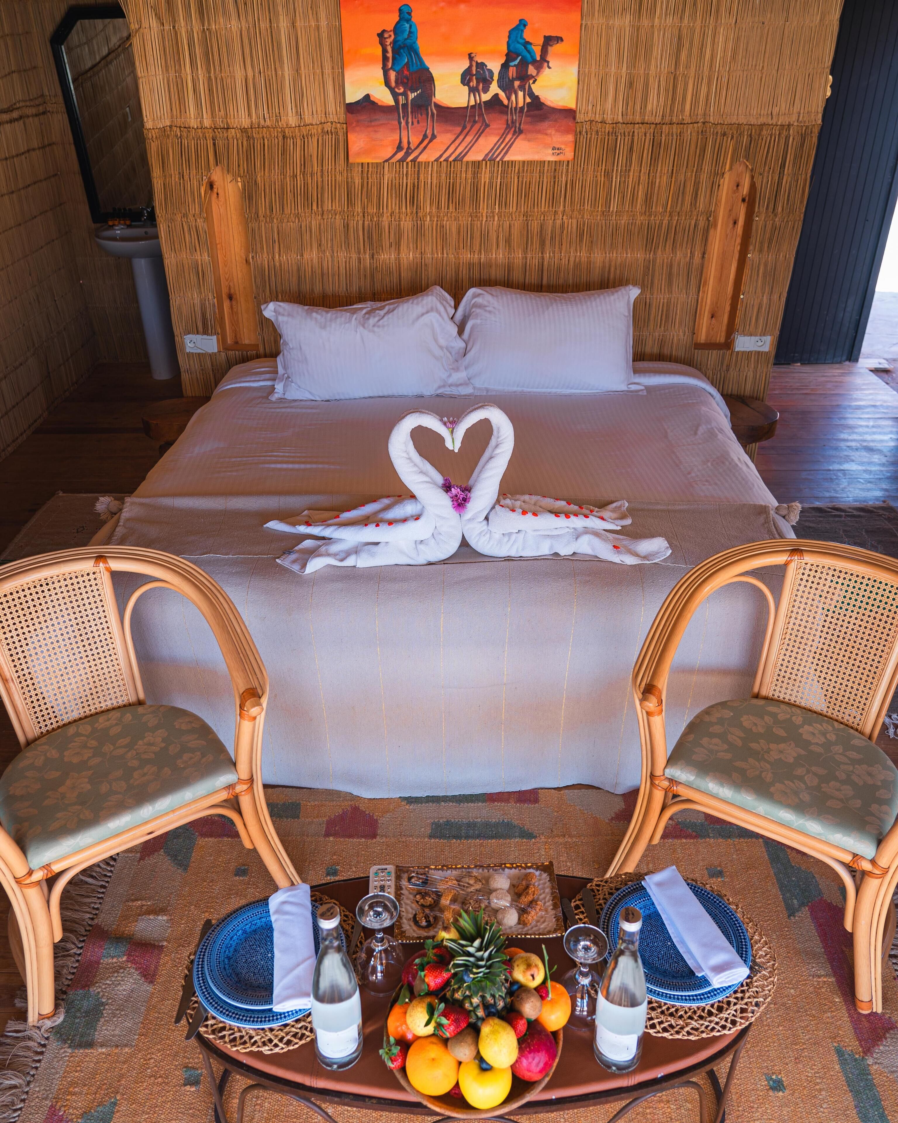 Une chambre d’hôtel avec un lit, deux fauteuils en osier, une petite table pour les fruits et le vin, et un tableau accroché au mur.