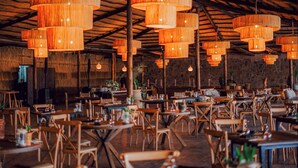 A spacious restaurant with multiple wooden tables and chairs, large hanging wooden chandeliers, and a rustic stone wall in the background. - Le Bedouin Agafay (Agafay)