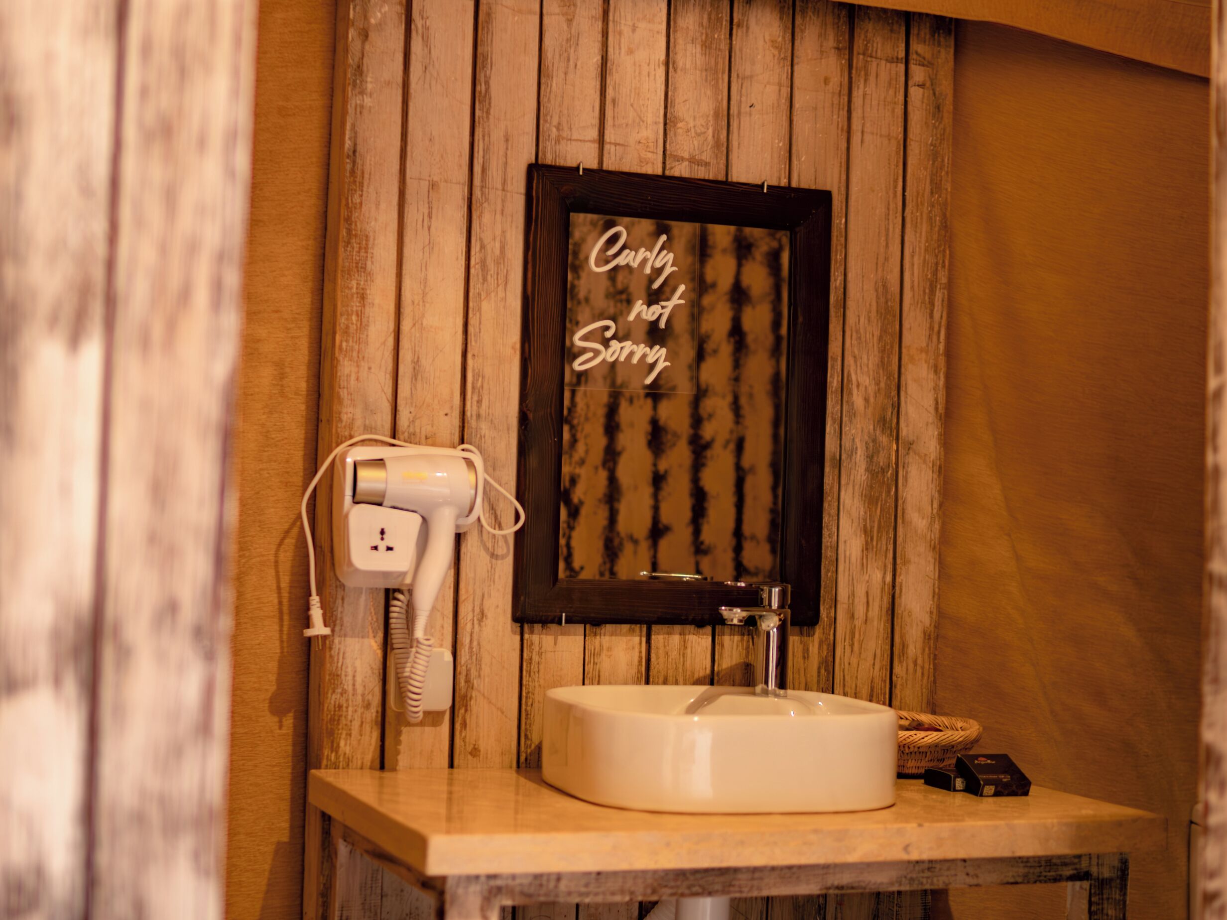 A bathroom with a wooden wall, a framed sign saying "Only hot water", a white sink, and a hairdryer plugged in.