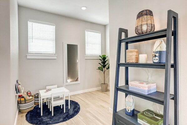 A room with a white table and chairs, a dark blue rug, a bookshelf with books and decorative items, and a potted plant.