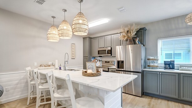 Una cocina moderna con armarios blancos, una isla central con sillas, iluminación colgante y electrodomésticos de acero inoxidable.