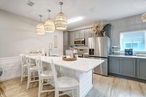 Una cocina moderna con armarios blancos, una isla central con sillas, iluminación colgante y electrodomésticos de acero inoxidable.