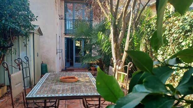 A patio with a tiled table and chairs, surrounded by greenery and a building in the background. - Small oasis in the center of Céret (Céret)