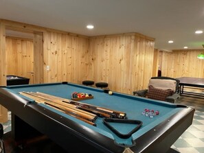 A pool table with pool cues and balls, a small seating area with a chair and ottoman, and wooden paneling on the walls.