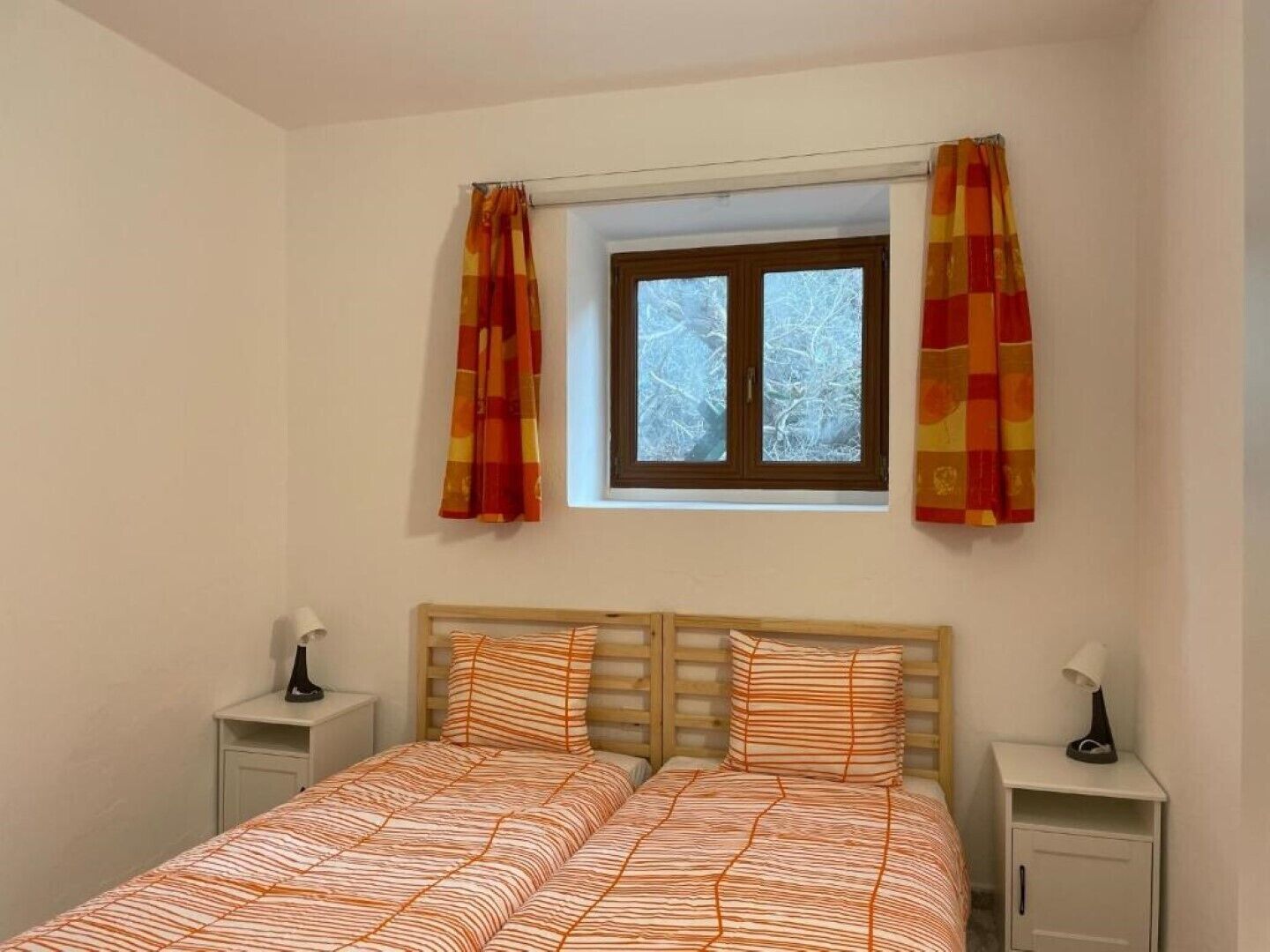 Un dormitorio con una cama, dos mesitas de noche, una ventana con cortinas y vistas a árboles.