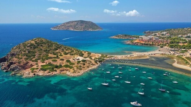 An aerial view of a coastal area with clear turquoise waters, a small island, and a beach with boats. - Platanus Gardenhouse (Saronikos)