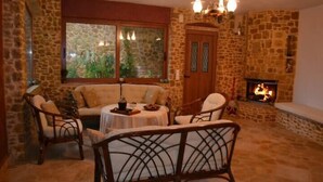 A cozy room with stone walls, a fireplace, a large window, and wicker furniture. - Platanus Gardenhouse (Saronikos)