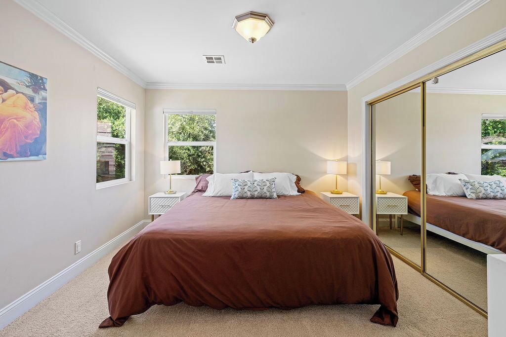 A bedroom with a large bed, two bedside tables with lamps, a large mirror, and a window with a view of greenery.