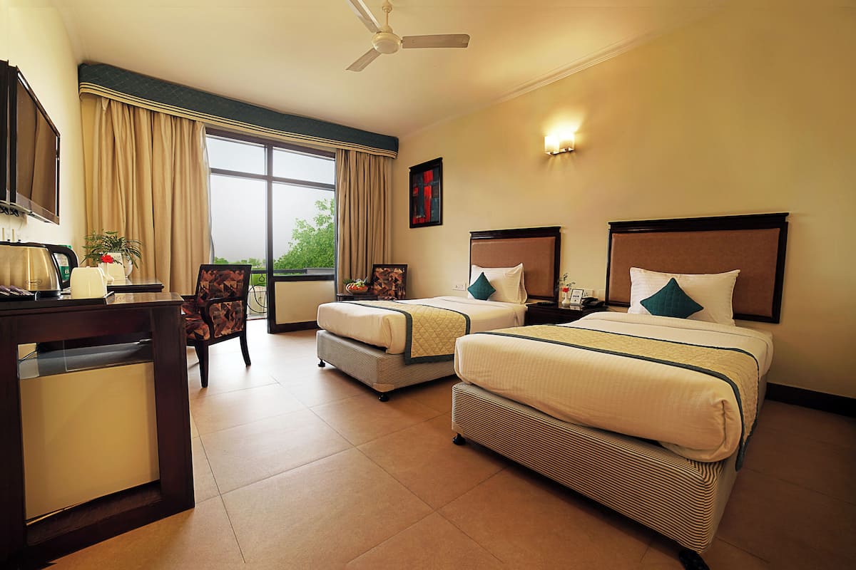 A hotel room with two beds, a desk, a chair, a ceiling fan, and a large window.
