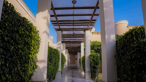 A covered walkway with a marble floor, flanked by white pillars and lush green hedges. - TreeHouse Sunrise Resort & Caravan Park (Neemrana)