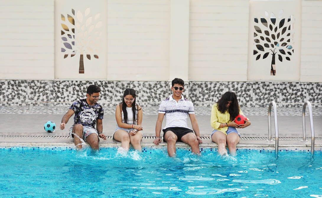 Una piscina con cuatro personas sentadas al borde, un muro de diseño azul y blanco, y siluetas de árboles decorativas.