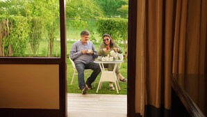 A man and a woman sitting at a small round table outdoors, with greenery and a fence in the background. - TreeHouse Sunrise Resort & Caravan Park (Neemrana)