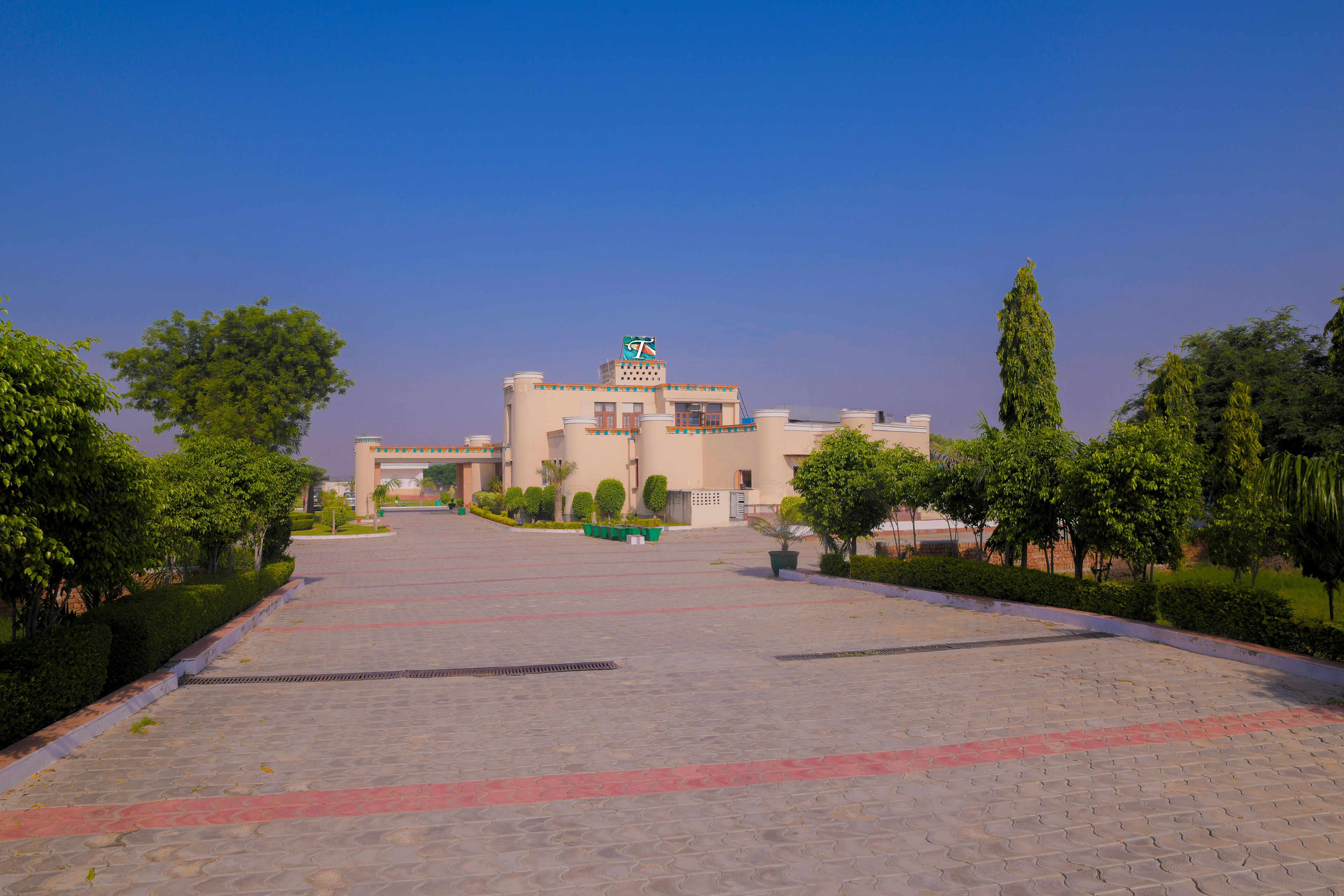 A spacious courtyard with a building featuring a prominent logo on the roof, surrounded by well-maintained greenery and a paved walkway.