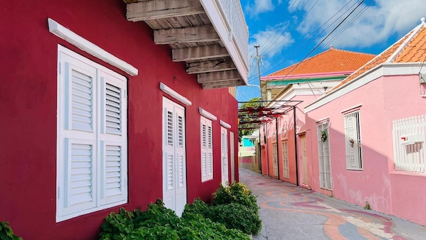 A narrow street lined with red and pink buildings, each with white shutters and a tiled roof. - Catherine Suites (Willemstad)
