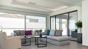 A modern living room with a sofa, armchair, and coffee table, large windows, and a view of the outside. - Balia Suites (Corfu)