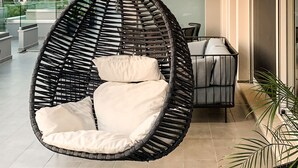 A modern outdoor seating area with a hanging chair and a sofa, a glass balustrade, and a view of the landscape. - Balia Suites (Corfu)