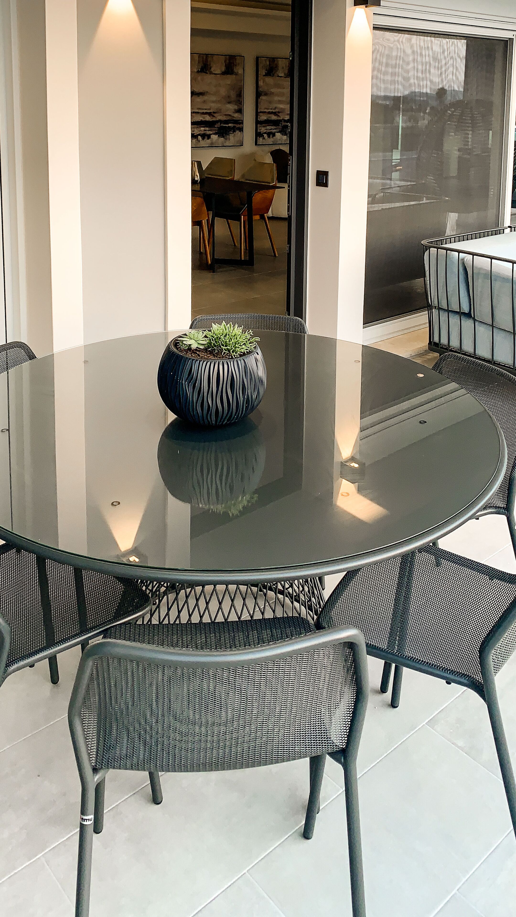 A round glass table with a textured black metal frame, surrounded by six black mesh chairs. A potted plant with a textured pot is placed on the table.