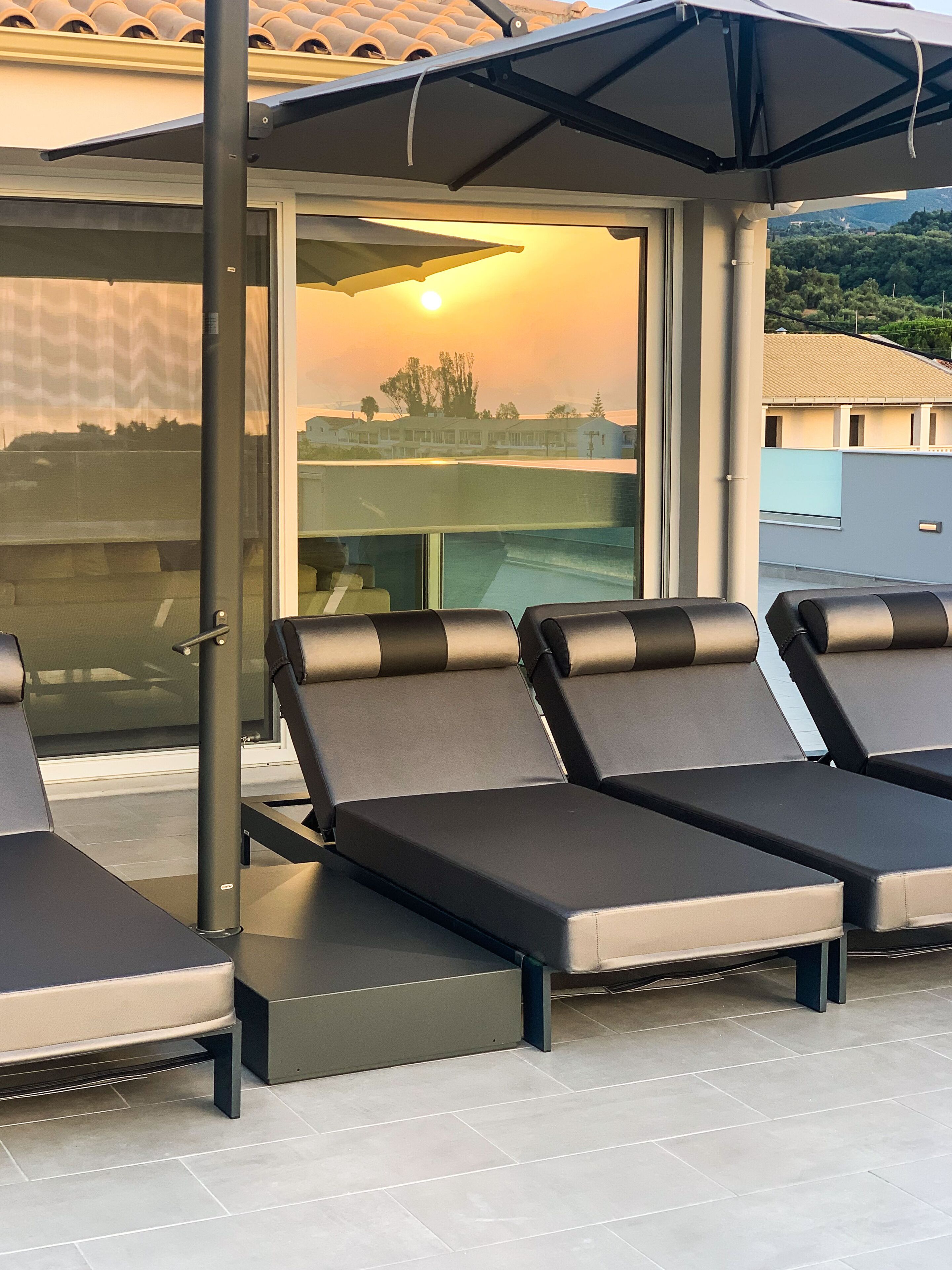 A rooftop terrace with four lounge chairs, a glass enclosure, and a view of a residential area during sunset.