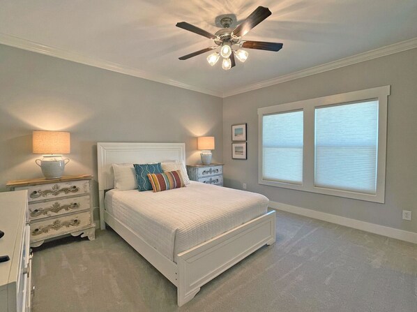 Quarto com cama, mesas de cabeceira, cômoda, ventilador de teto e duas janelas com persianas.