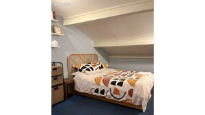 Um quarto com uma cama, uma mesinha de cabeceira, um cesto de vime e uma estante com livros e um vaso.